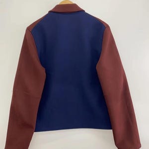 May include: A color-blocked jacket featuring a solid navy blue back panel and sleeves in a deep maroon shade. The garment includes a matching maroon collar and is displayed on a wooden hanger, showcasing a clean, structured design with long sleeves.