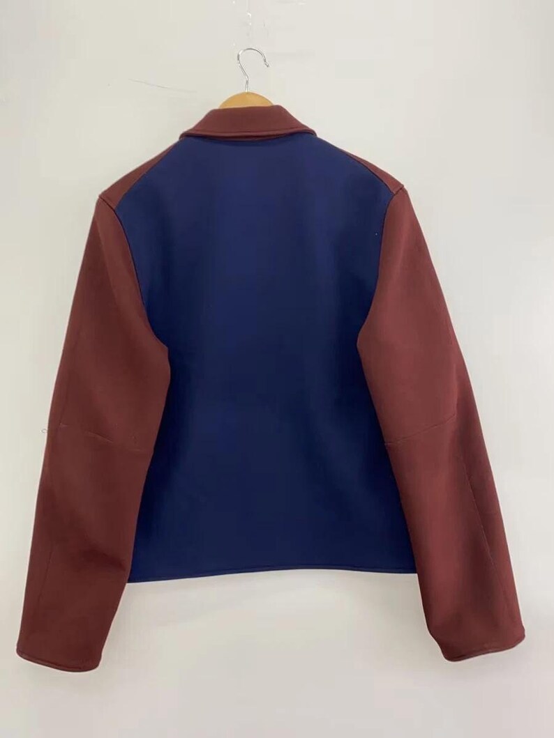 May include: A color-blocked jacket featuring a solid navy blue back panel and sleeves in a deep maroon shade. The garment includes a matching maroon collar and is displayed on a wooden hanger, showcasing a clean, structured design with long sleeves.