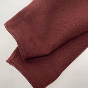 May include: A close-up view of the sleeves of a dark burgundy garment. The fabric features a distinct, fine diagonal twill weave texture. The cuffs are finished with clean, straight stitching along the edges, showing the deep red color.