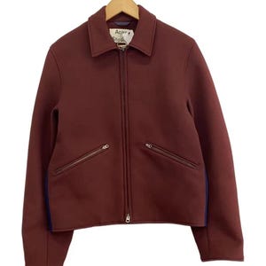 May include: A burgundy zip-up jacket with a classic collar, long sleeves, and two slanted zippered pockets on the front. The garment features a visible Acne Studios brand label at the neckline and dark blue side panels along the torso.