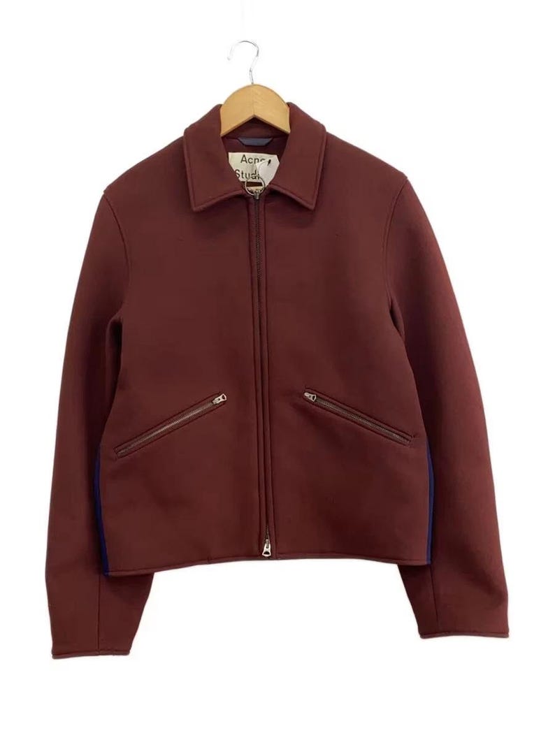 May include: A burgundy zip-up jacket with a classic collar, long sleeves, and two slanted zippered pockets on the front. The garment features a visible Acne Studios brand label at the neckline and dark blue side panels along the torso.