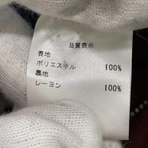 May include: A white fabric garment care label with black Japanese text. The label indicates the outer material is 100% polyester and the lining material is 100% rayon. The text includes the header Quality Indication, followed by Polyester 100% and Rayon 100%.