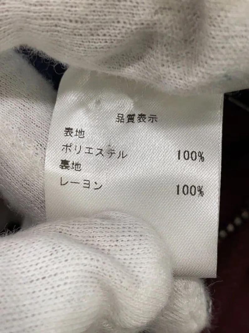 May include: A white fabric garment care label with black Japanese text. The label indicates the outer material is 100% polyester and the lining material is 100% rayon. The text includes the header Quality Indication, followed by Polyester 100% and Rayon 100%.