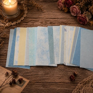 May include: A collection of rectangular paper sheets in various shades of blue, light blue, and pale yellow, featuring diverse textures and patterns, arranged in a fan-like display on a wooden surface with dried flowers and a candle.