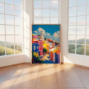 Può includere: Un dipinto rettangolare verticale che raffigura una città collinare con edifici colorati nelle tonalità del blu, rosa, giallo e arancione sotto un cielo azzurro brillante con nuvole bianche. Un alto campanile spicca tra le case.