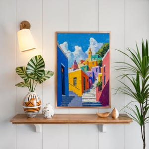 Peut inclure: Un tableau encadré représentant un village méditerranéen aux couleurs vives avec des bâtiments jaunes, orange et roses, un escalier en pierre et un ciel bleu vif avec des nuages blancs, posé sur une étagère en bois avec une plante.