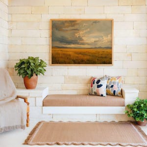 Puede incluir: Un cuadro rectangular de paisaje con un campo dorado bajo un cielo nublado y dramático, colocado sobre un banco empotrado con dos cojines estampados, una alfombra tejida y dos plantas verdes en una habitación con paredes de piedra.
