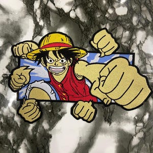 May include: An embroidered patch featuring a cartoon character with black hair, a yellow straw hat, and a red vest. The character is depicted with multiple arms extended forward in a punching motion against a rectangular blue background with white clouds.