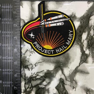 May include: A circular embroidered patch featuring a black background with a white, orange, and grey rocket ship graphic. The text PROJECT HAIL MARY is stitched in white along the bottom. A ruler shows the patch is approximately 4 inches wide.