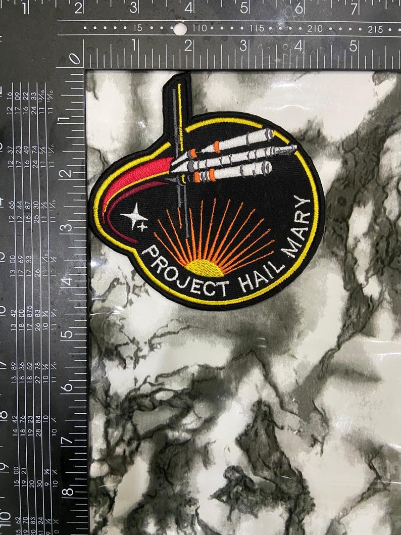 May include: A circular embroidered patch featuring a black background with a white, orange, and grey rocket ship graphic. The text PROJECT HAIL MARY is stitched in white along the bottom. A ruler shows the patch is approximately 4 inches wide.