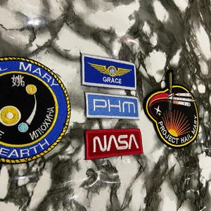 May include: A collection of five embroidered patches including a circular blue and black patch with text, a rectangular blue patch with yellow wings and the text GRACE, a light blue PHM patch, a red NASA patch, and a black Project Hail Mary patch.