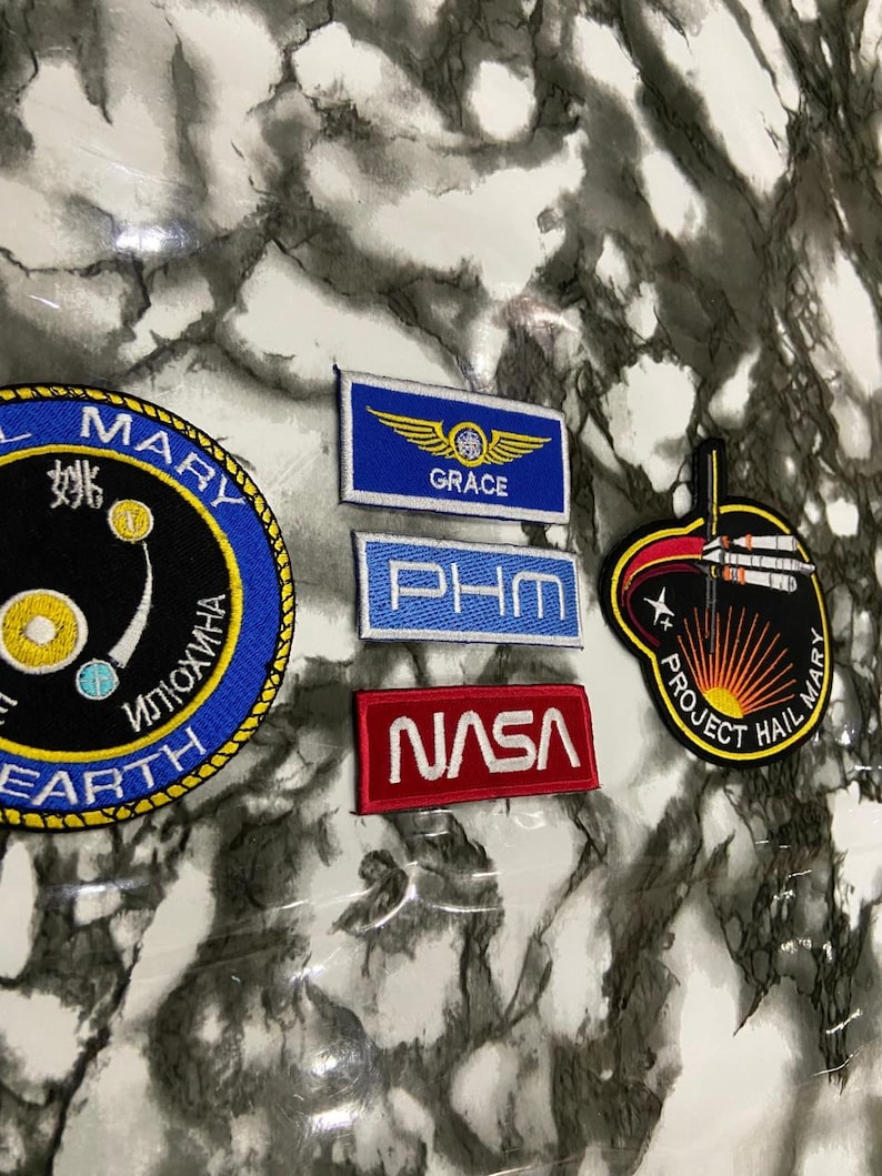 May include: A collection of five embroidered patches including a circular blue and black patch with text, a rectangular blue patch with yellow wings and the text GRACE, a light blue PHM patch, a red NASA patch, and a black Project Hail Mary patch.