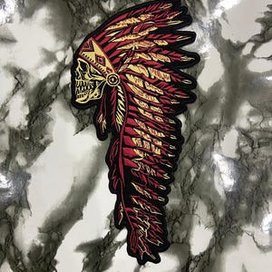 May include: An embroidered patch featuring a skull wearing a traditional-style feathered headdress. The design uses a colour palette of deep red, black, and gold thread, with intricate stitching detailing the skull's features and the individual feathers.