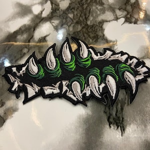 May include: An embroidered patch featuring a series of sharp, white, claw-like shapes emerging from a black background with vibrant green accents. The design has a jagged, ripped edge effect with black stitching outlining the entire shape.