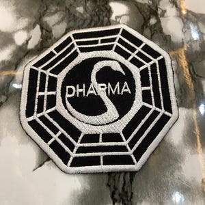 May include: An octagonal embroidered patch featuring a black and white geometric design. The center displays a white swan silhouette intertwined with the letter S, and the text DHARMA is stitched in white across the center of the swan graphic.