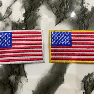 May include: Two rectangular embroidered patches featuring the United States flag design. The left patch has a silver border, while the right patch has a gold border. Both patches display a blue canton with white stars and alternating red and white stripes.