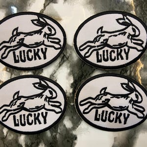 May include: Four oval-shaped embroidered patches featuring a black and white graphic of a running rabbit. Each patch has a black border and the word LUCKY printed in black capital letters beneath the rabbit illustration on a white background.