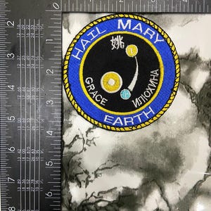 May include: A circular embroidered patch with a black center and a blue border featuring the text "HAIL MARY GRACE EARTH" and Cyrillic characters. The center displays a yellow circle, a small blue circle, and a white character. A ruler shows a 3-inch diameter.