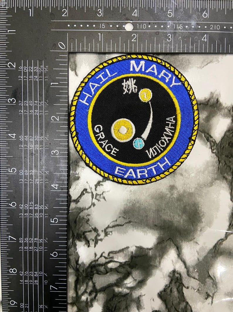 May include: A circular embroidered patch with a black center and a blue border featuring the text "HAIL MARY GRACE EARTH" and Cyrillic characters. The center displays a yellow circle, a small blue circle, and a white character. A ruler shows a 3-inch diameter.
