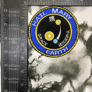 May include: A circular embroidered patch with a blue border and yellow rope-like trim. The center is black with white text reading HAIL MARY, GRACE, and EARTH, plus Cyrillic text. It features yellow and blue circular icons and measures 3 inches.