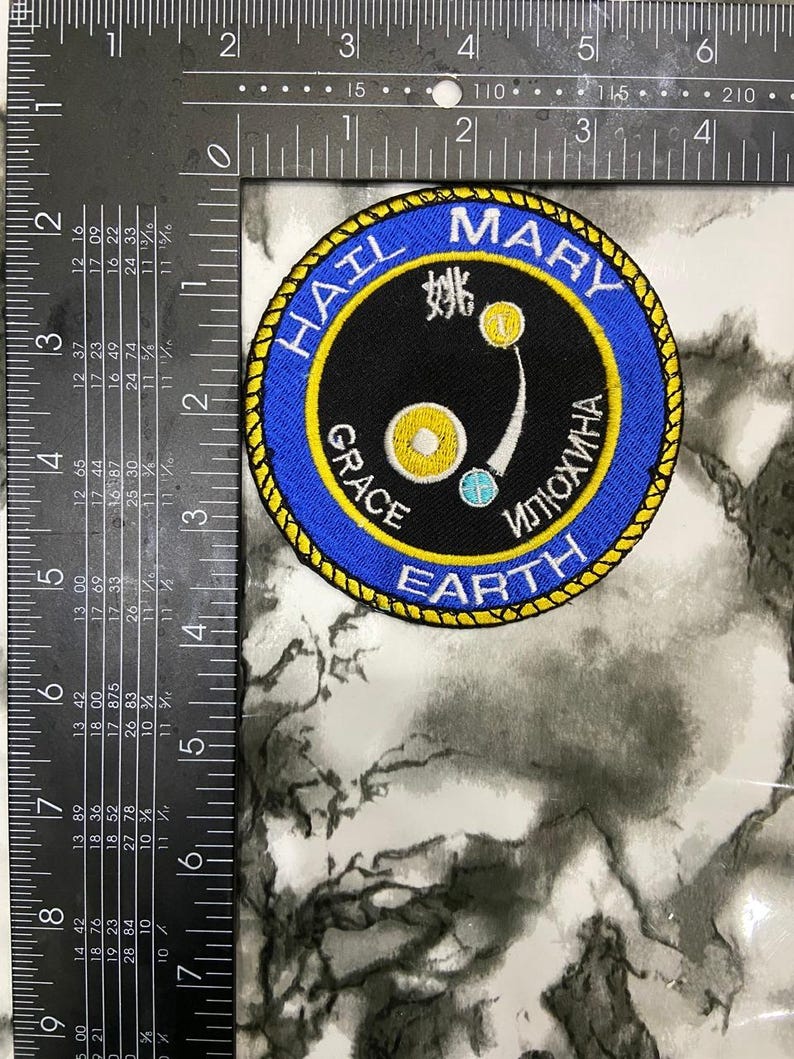 May include: A circular embroidered patch with a blue border and yellow rope-like trim. The center is black with white text reading HAIL MARY, GRACE, and EARTH, plus Cyrillic text. It features yellow and blue circular icons and measures 3 inches.