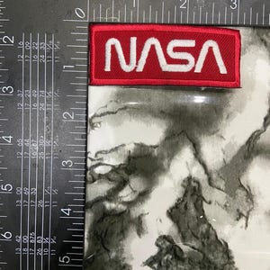 May include: A rectangular embroidered patch featuring the white NASA logo on a red background. The patch is positioned against a black metal framing square, showing a width of approximately 3.5 inches and a height of approximately 1.25 inches.