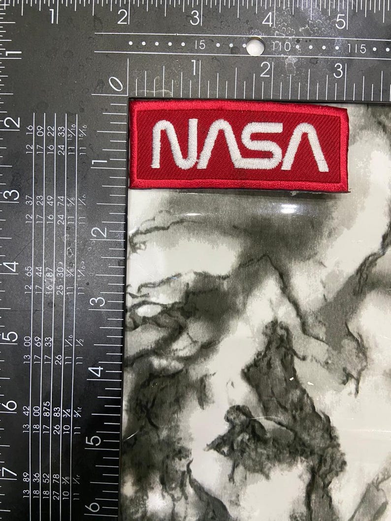 May include: A rectangular embroidered patch featuring the white NASA logo on a red background. The patch is positioned against a black metal framing square, showing a width of approximately 3.5 inches and a height of approximately 1.25 inches.