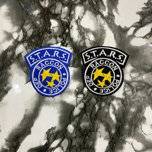 Puede incluir: Dos parches bordados con un diseño circular con tres estrellas amarillas en el centro. El texto S.T.A.R.S. aparece en una banda arqueada arriba, con RACCON POLICE DEP escrito alrededor del borde. Un parche es azul y el otro es negro.
