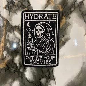 May include: A rectangular embroidered patch with a black background and white stitching. It features a hooded skeleton holding a water bottle, a crescent moon, and stars. The text reads HYDRATE at the top and OUTLIVE YOUR ENEMIES at the bottom.