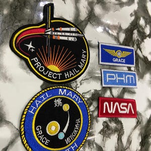 May include: A collection of embroidered patches including two circular mission-style emblems featuring space imagery and text like Project Hail Mary, Hail Mary, and Earth, alongside three rectangular patches labeled Grace, PHM, and NASA in red, blue, and white.
