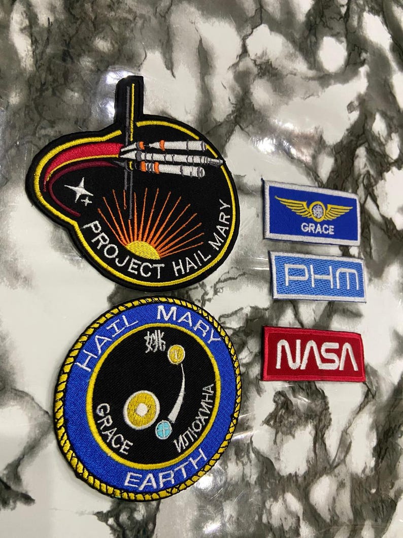 May include: A collection of embroidered patches including two circular mission-style emblems featuring space imagery and text like Project Hail Mary, Hail Mary, and Earth, alongside three rectangular patches labeled Grace, PHM, and NASA in red, blue, and white.