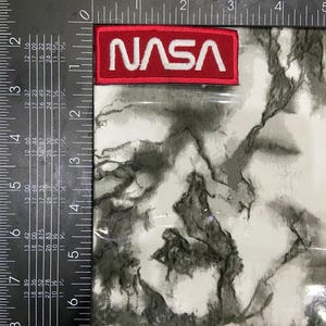 May include: A rectangular NASA patch with a red background and white embroidered lettering. The patch is positioned next to a black metal framing square with white imperial measurement markings, showing the patch is 3 inches wide.