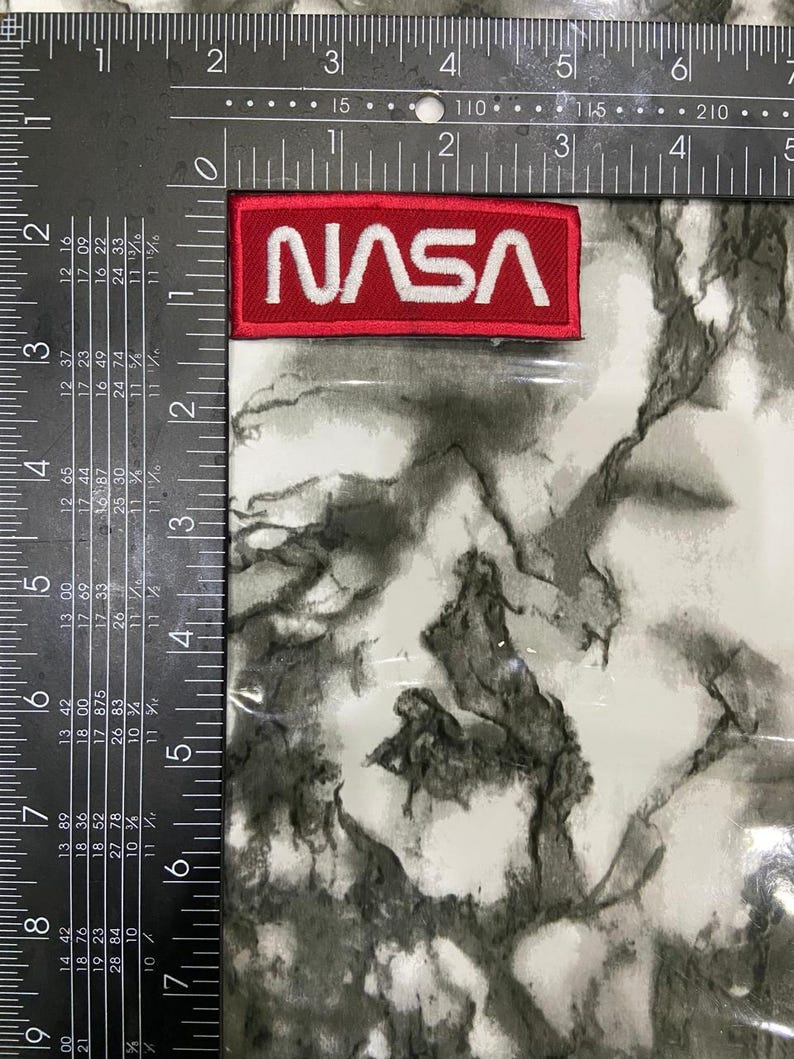 May include: A rectangular NASA patch with a red background and white embroidered lettering. The patch is positioned next to a black metal framing square with white imperial measurement markings, showing the patch is 3 inches wide.
