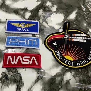 May include: Four embroidered patches including a blue rectangular patch with yellow wings and the text GRACE, a light blue rectangular patch with the text PHM, a red rectangular patch with the text NASA, and a black circular patch with a rocket and text.