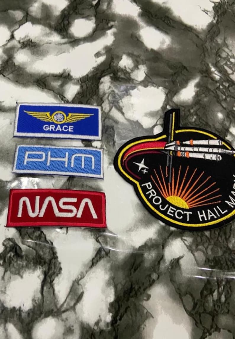 May include: Four embroidered patches including a blue rectangular patch with yellow wings and the text GRACE, a light blue rectangular patch with the text PHM, a red rectangular patch with the text NASA, and a black circular patch with a rocket and text.