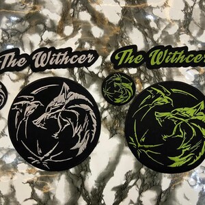 May include: Two sets of embroidered patches featuring a wolf head emblem and the text The Withcer. One set is black with white embroidery, and the other is black with lime green embroidery. Each set includes one large circular patch and one small circular patch.