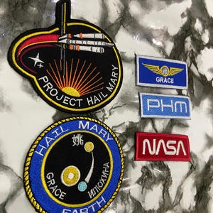 May include: A collection of five embroidered patches featuring space-themed designs. Two are circular, one with a black background and Project Hail Mary text, the other blue with Earth and Grace text. Three are rectangular with Grace, PHM, and NASA text.