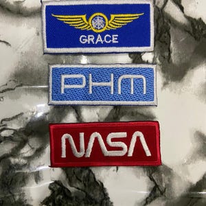 May include: Three rectangular embroidered patches. The top patch is blue with yellow wings, a central emblem, and the text GRACE. The middle patch is light blue with the white text PHM. The bottom patch is red with the white text NASA.