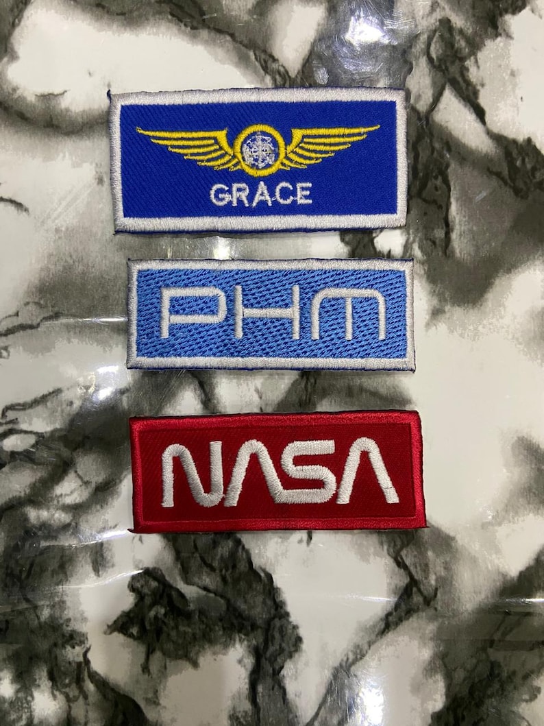 May include: Three rectangular embroidered patches. The top patch is blue with yellow wings, a central emblem, and the text GRACE. The middle patch is light blue with the white text PHM. The bottom patch is red with the white text NASA.