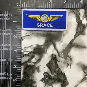 May include: A rectangular blue embroidered patch featuring a yellow winged emblem with a central circular design and the name GRACE in white capital letters. The patch is positioned against a metal ruler showing a width of approximately 3.5 inches.