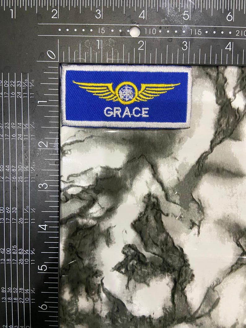 May include: A rectangular blue embroidered patch featuring a yellow winged emblem with a central circular design and the name GRACE in white capital letters. The patch is positioned against a metal ruler showing a width of approximately 3.5 inches.