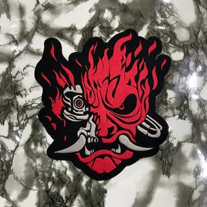 May include: An embroidered patch featuring a stylized red and black oni mask design with flames rising from the top. The mask includes mechanical details on one side, sharp white fangs, and a black border, creating a bold graphic appearance.