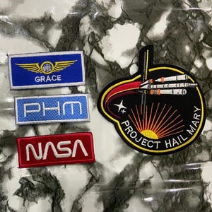 May include: Four embroidered patches: a blue rectangular patch with gold wings and the text GRACE, a light blue rectangular patch with the text PHM, a red rectangular patch with the white text NASA, and a large circular black patch featuring a rocket.