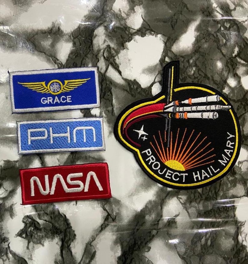 May include: Four embroidered patches: a blue rectangular patch with gold wings and the text GRACE, a light blue rectangular patch with the text PHM, a red rectangular patch with the white text NASA, and a large circular black patch featuring a rocket.