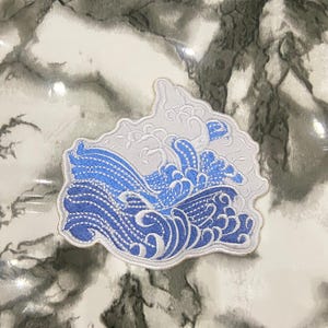 Peut inclure: Un écusson brodé représentant un motif de vague océanique stylisée avec des fils bleu clair et bleu foncé sur un fond blanc. L'écusson présente une forme irrégulière distincte qui suit les contours du motif de la vague déferlante.