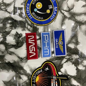 May include: A collection of five embroidered patches. One circular patch features a blue border, black center, and text reading HAIL MARY, EARTH, and Russian characters. Three rectangular patches include NASA in red, PHM in light blue, and GRACE with wings in blue. One large circular patch depicts a rocket and the text PROJECT HAIL MARY.