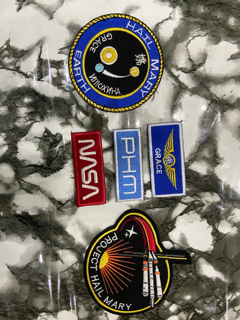 May include: A collection of five embroidered patches. One circular patch features a blue border, black center, and text reading HAIL MARY, EARTH, and Russian characters. Three rectangular patches include NASA in red, PHM in light blue, and GRACE with wings in blue. One large circular patch depicts a rocket and the text PROJECT HAIL MARY.