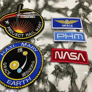 May include: A collection of five embroidered patches featuring space-themed designs. Two are circular, one with a rocket and 'PROJECT HAIL MARY' text, another with celestial bodies and 'HAIL MARY GRACE EARTH' text. Three are rectangular with 'GRACE', 'PHM', and 'NASA' text.