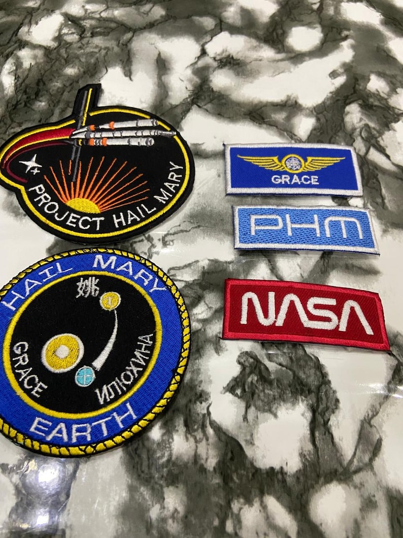 May include: A collection of five embroidered patches featuring space-themed designs. Two are circular, one with a rocket and 'PROJECT HAIL MARY' text, another with celestial bodies and 'HAIL MARY GRACE EARTH' text. Three are rectangular with 'GRACE', 'PHM', and 'NASA' text.