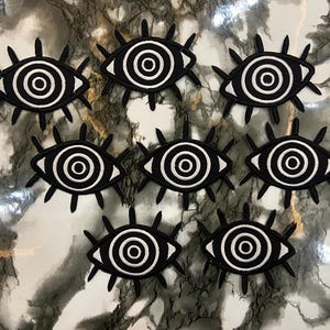 Pode incluir: Oito emblemas bordados em forma de olho, cada um apresentando um design de círculos concêntricos em preto e branco para a íris e a pupila, rodeados por cílios pretos que irradiam para fora a partir do contorno amendoado do olho.